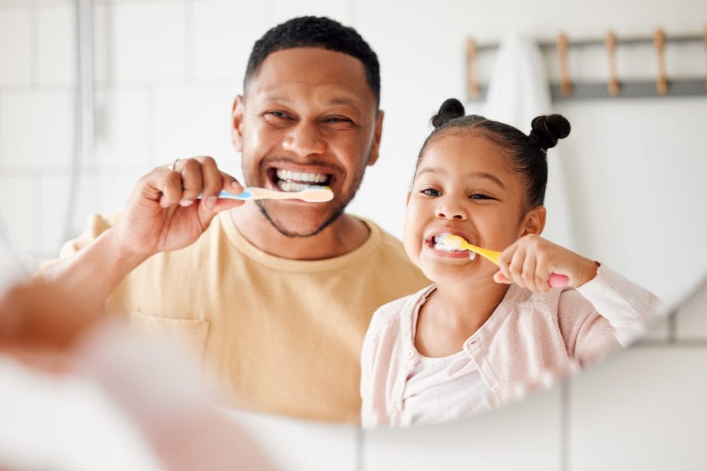 Smiling dad and young daughter brushing their teeth together in front of bathroom mirror – Novato Dentist 